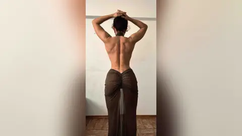 Muscular woman in a brown backless maxi dress posing in a minimalist room, showcasing a toned back and physique.
