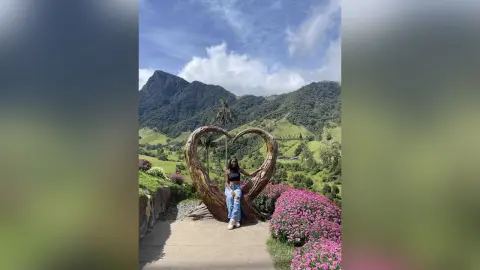 NatyGlow sits inside a large heart-shaped wooden frame overlooking a lush, green mountain valley under a blue sky.