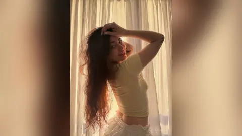 Young woman in a yellow t-shirt posing by a sunlit window in a warm, dreamy indoor setting.