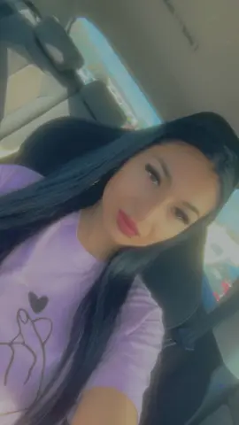 Smiling young woman with long dark hair wearing a lavender graphic t-shirt sitting in a car.