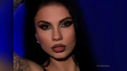CutemEmma poses with long dark hair and heavy eye makeup against a deep blue background, looking directly at the camera.