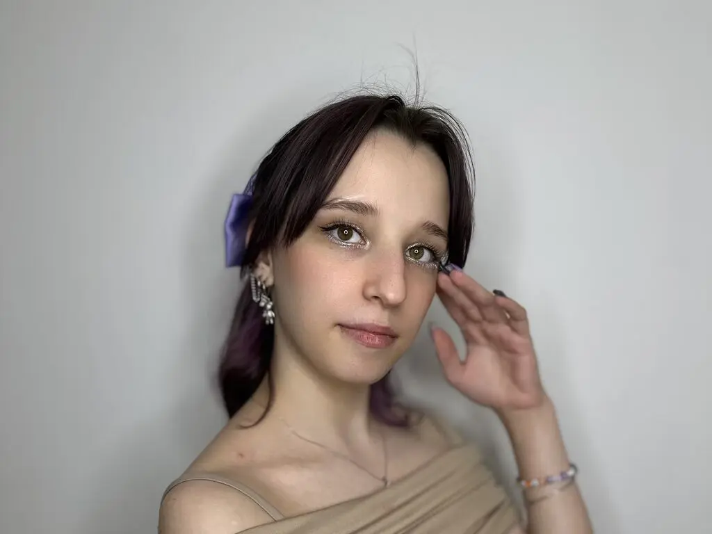 Young woman with glitter eyeshadow and purple hair bow wearing a beige top against a plain white background.