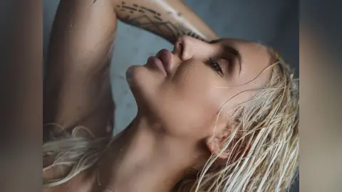 Anita poses with her eyes closed and wet blonde hair, tilting her head back while resting her arm against her head.