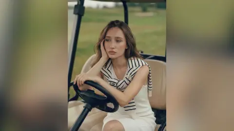 Melancholic woman in a striped sleeveless top sitting in a golf cart on a grassy field.