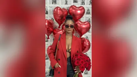 Stylish woman in a red blazer holding a rose bouquet against a backdrop of red heart balloons.