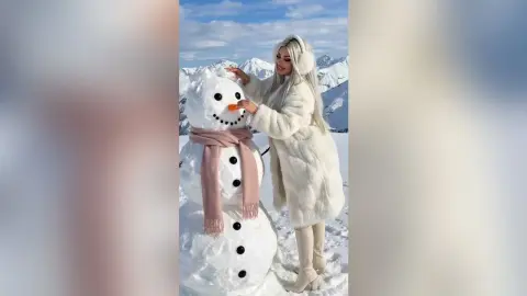 Blonde woman in a white faux fur coat smiling next to a large snowman in a sunny snowy mountain landscape.
