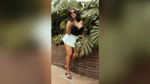 Stylish woman in a black corset and white mini skirt taking a selfie amidst lush green tropical plants.