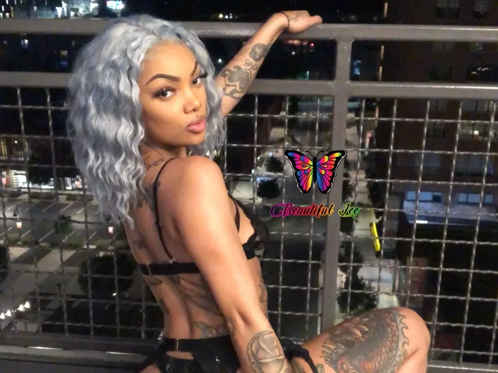 Tattooed woman with wavy silver hair in a black bikini posing on a city balcony at night.