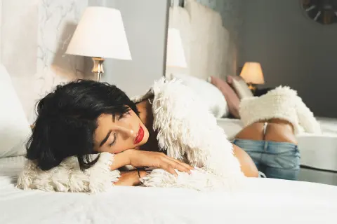 Young woman in a white fuzzy sweater relaxing on a bed in a cozy, bright bedroom setting.