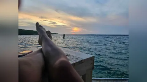 Relaxed view of bare feet resting on a wooden deck overlooking a calm ocean at sunset.