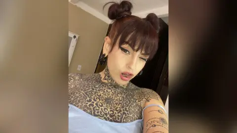 Tattooed woman with dark bangs and buns wearing a blue tank top in a casual indoor setting.