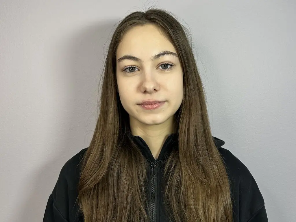 Young woman with long brown hair wearing a black zip-up hoodie against a neutral grey wall background.