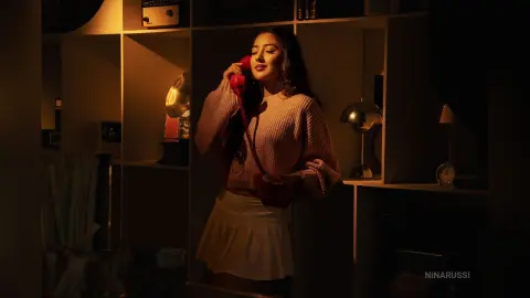 Young woman in a pink sweater and white skirt talking on a red vintage telephone in warm, moody indoor lighting.