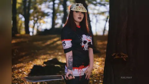 Woman in dark cosplay outfit holding a large sword in a moody autumn forest setting.