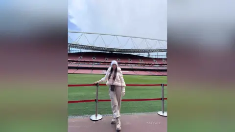 Young woman in a white puffer jacket and beanie standing at a large outdoor sports stadium on a cloudy day.
