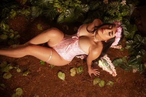 Sensual woman in pink lace lingerie reclining on forest floor surrounded by lush green tropical leaves.