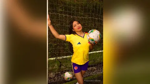 NairaWestbrook poses in a yellow soccer jersey and purple shorts, holding a soccer ball in front of a net.