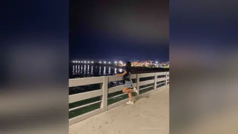 Young woman in a black top and denim shorts standing on a pier by the ocean at night under city lights.