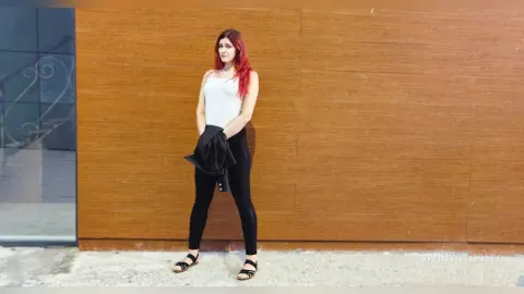 Red-haired woman in a white tank top and black leggings standing against a wooden wall background.
