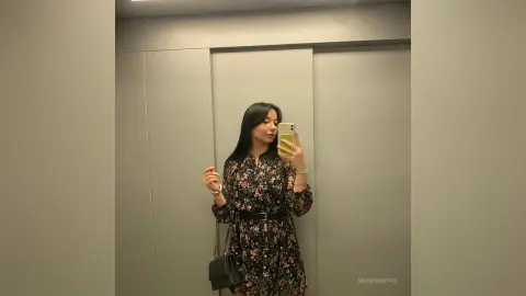 Young woman in a floral black midi dress taking a mirror selfie in a modern minimalist hallway.