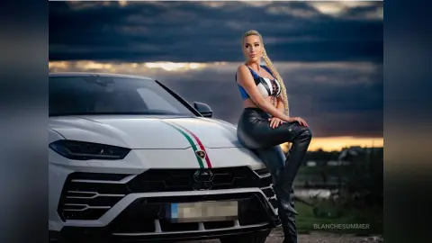 Stylish blonde woman in a blue crop top and black leather pants leaning on a white luxury SUV at sunset.
