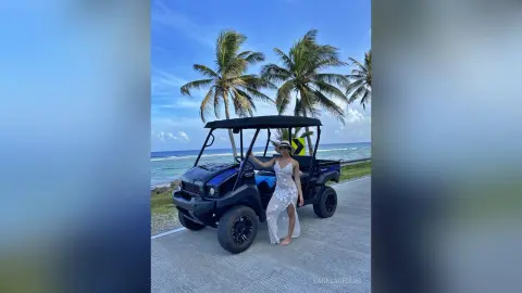 Woman in a white summer dress standing by a golf cart on a sunny tropical beach road.