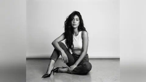 Elegant woman in a white tank top and jeans sitting on a studio floor in a moody black and white portrait.