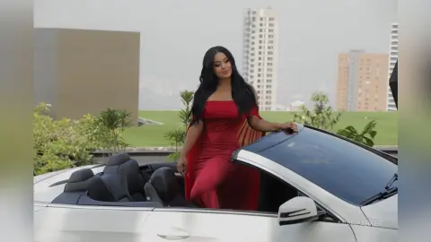 Miah poses in a red dress while sitting in a white convertible car outdoors.