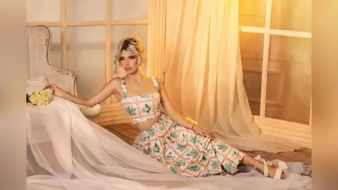 Blonde woman in floral crop top and skirt sitting on a white bed by a sunlit window in a dreamy aesthetic mood.
