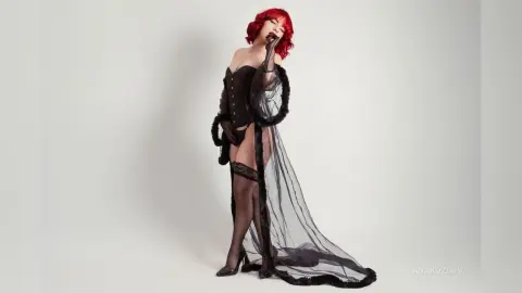 Elegant woman with red hair in a black corset and sheer lace gown posing against a white studio background.