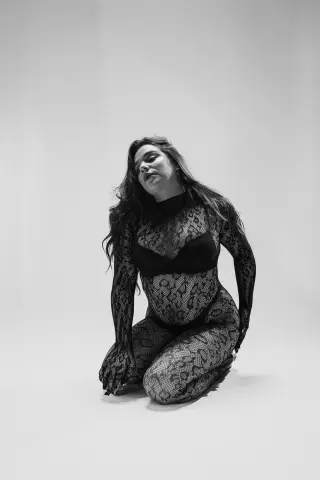 Curvy woman in a black lace bodysuit posing elegantly in a minimalist studio setting.
