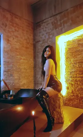 Glamorous woman in a brown leather dress and black thigh-high boots sitting in a moody, rustic brick room.