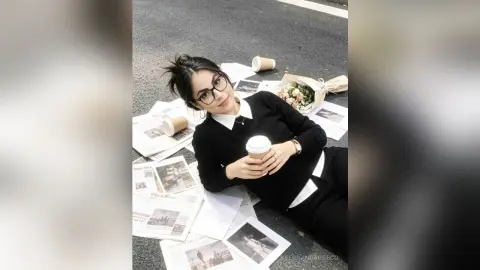 Young woman in a black sweater relaxing on newspapers with coffee and flowers on a city street.