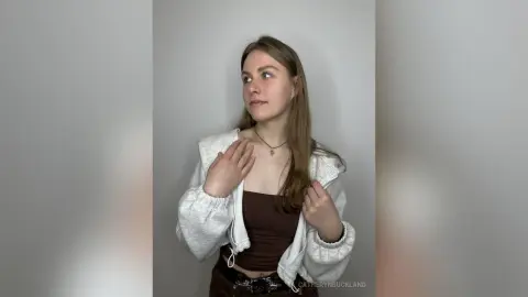 CatherynBuckland wearing a white zip-up hoodie and a brown tank top, looking off to the side against a plain grey background.