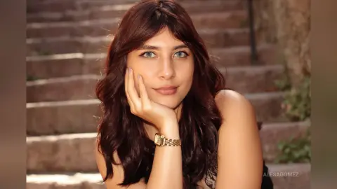 Smiling woman with long wavy brown hair wearing a gold watch sitting on stone stairs in warm sunlight.