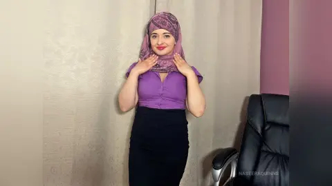 Smiling woman in a purple blouse and black skirt wearing a patterned mauve hijab in a bright indoor setting.