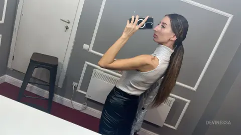 DeviNessa holds a smartphone up to a mirror, wearing a white sleeveless turtleneck and black leather pants.