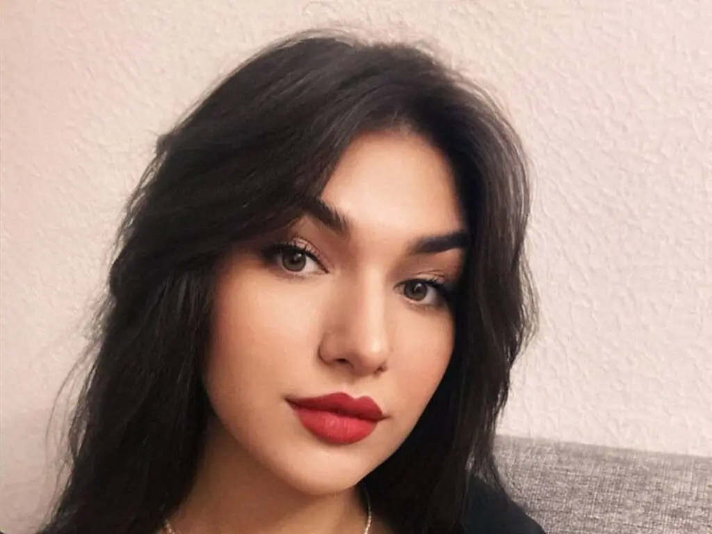 Young woman with long dark hair and red lipstick posing for a close-up portrait against a neutral background.