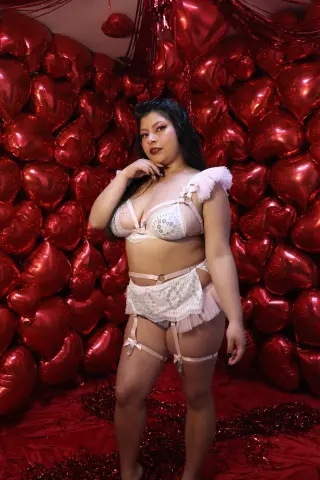 Woman in white lace lingerie posing against a romantic red heart balloon backdrop.