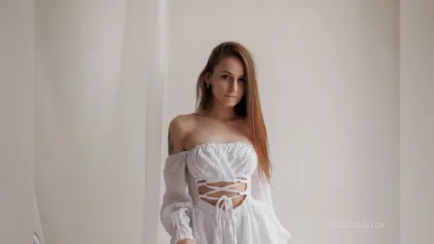 IngridBeckton poses in a white off-the-shoulder lace top with a lace-up front, showing her long brown hair and shoulder tattoo against a white wall.