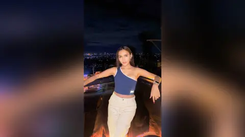 Stylish woman in a blue halter top and white pants posing on a rooftop at night with city lights background.