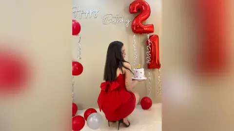 Young woman in a red dress holding a birthday cake near red balloons in a festive celebratory setting.