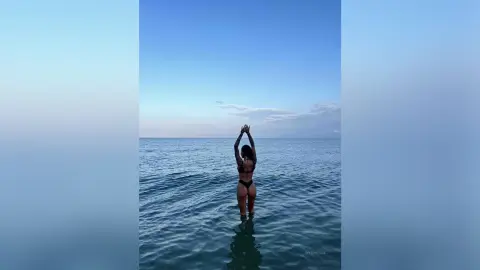 Woman in black bikini standing in calm ocean water under a serene blue sky at sunset.