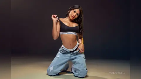 Young woman in black Calvin Klein bralette and baggy denim jeans posing confidently against a dark studio background.