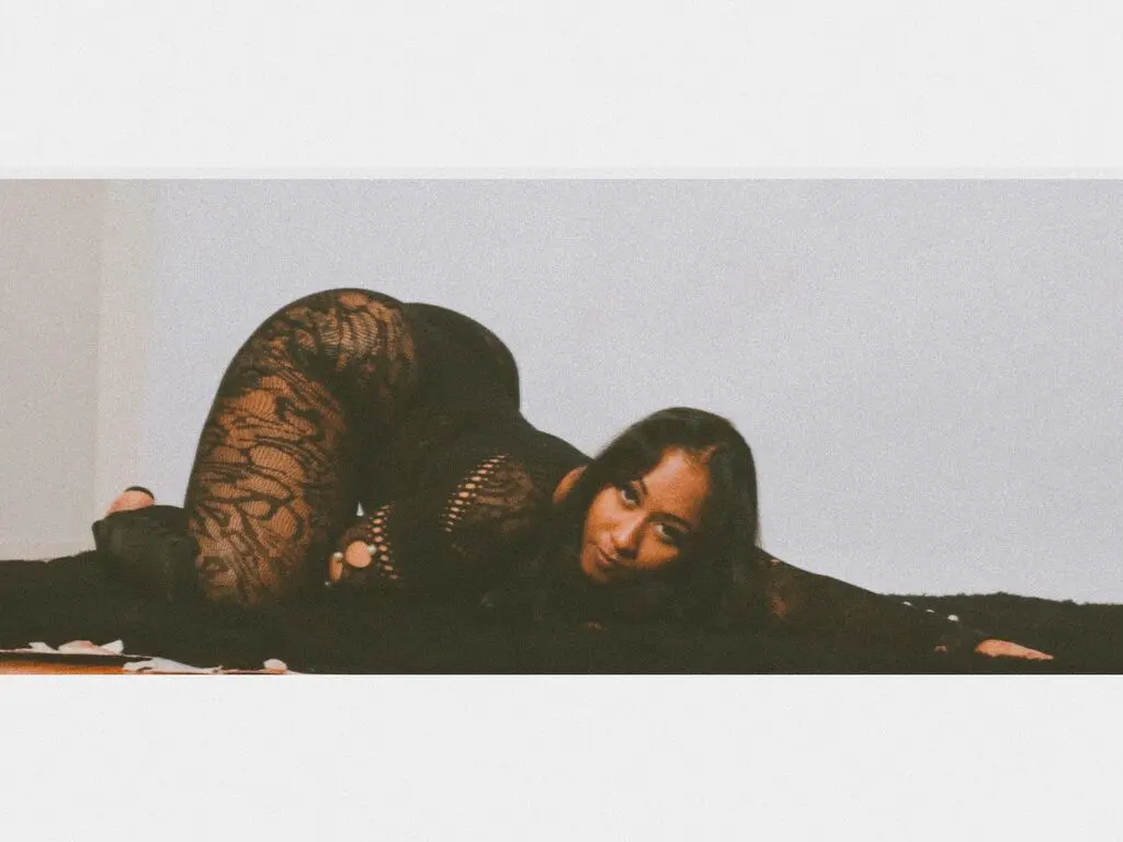 Curvy woman in black lace bodysuit lounging on a dark rug in a minimalist room with a moody aesthetic.