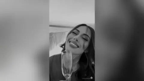 Smiling young woman in a dark top holding a champagne flute in a bright, celebratory indoor setting.