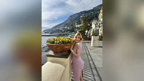 Blonde woman in a pink ribbed dress standing by yellow flowers on a sunny coastal terrace overlooking the sea.