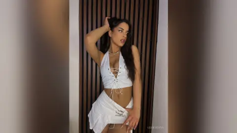 Young woman in a white lace halter top and skirt posing confidently against a modern wood slat wall.