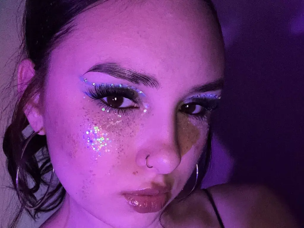 Young woman with glittery festival makeup and blue eyeliner under moody purple neon lighting.