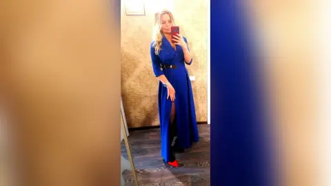MilenaBia stands in front of a patterned wall wearing a long blue dress with a slit and a black belt.